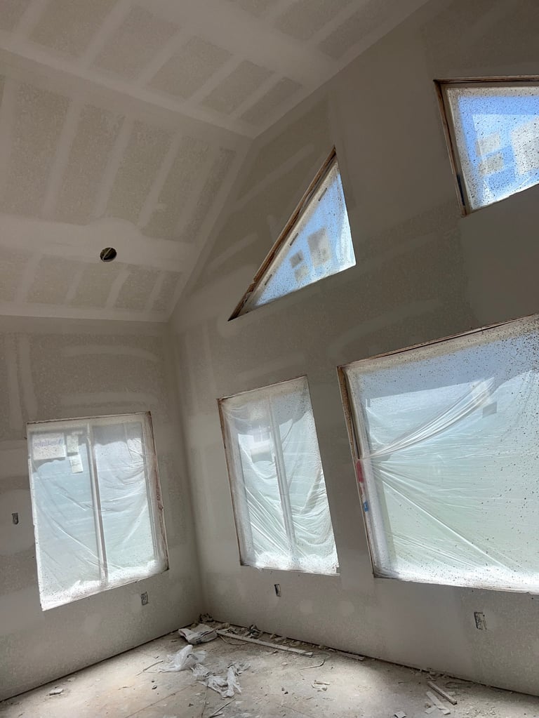 Interior of unfinished room with drywall walls, beamed ceiling, and multiple windows covered with plastic sheeting during construction
