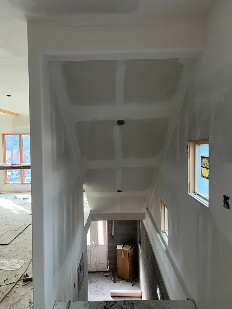 Interior hallway of a residential construction site with drywall walls and ceiling, wooden windows, and cardboard boxes on the floor